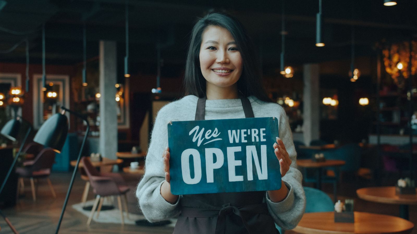 A cafe owner in an apron holding a 'Yes we're open' sign